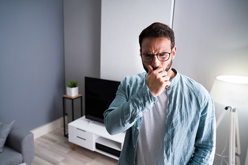 A man wearing glasses and a light blue button-up shirt over a grey t-shirt stands in a modern living room while pinching his nose with his hand, reacting with a look of intense disgust or discomfort.