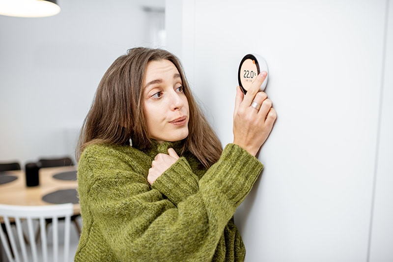 woman wearing a sweater, shivering as she turns up her thermostat.