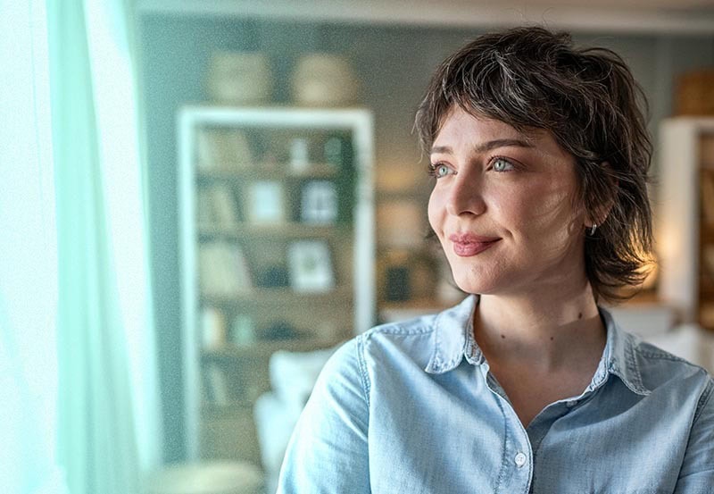 A young woman with short dark hair and blue eyes wearing a light blue denim shirt, smiling softly as she gazes out a window in her home.
